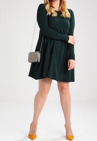 Ciemnozielona sukienka z długim rękawem i rozkloszowaną spódnicą, zestawiona z srebrną torebką na ramię i musztardowymi szpilkami. Gładki materiał, długość do kolan.