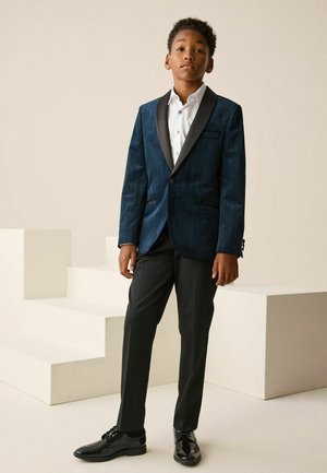 Tmavě modrý sametový saka s černými saténovými klopami, doplněné bílou košilí a černými společenskými kalhotami. Lesklé černé boty podtrhují celý vzhled.