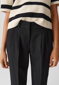 Haut en tricot avec des rayures horizontales crème et noir, associé à un pantalon noir cintré avec des plis et une fermeture éclair à l'avant.
