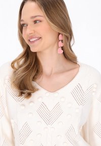 Cremefarbener Strickpullover mit strukturierten Mustern und Bollen; rosa hängende Herzohrringe. Sanfte Farben und ein V-Ausschnitt-Design.