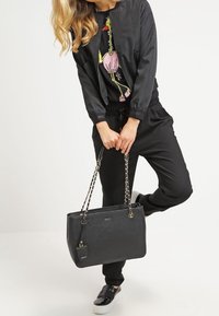 Czarna torba na ramię z łańcuszkowymi paskami i małą metką, teksturowana powierzchnia, prostokątny kształt, charakteryzująca się nowoczesnym wzornictwem.