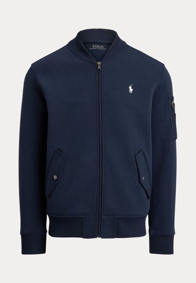 Blouson bomber bleu marine zippé avec col, poignets et bas côtelés, poches à rabat avec boutons, poche zippée sur la manche et logo Polo Ralph Lauren blanc sur la poitrine.