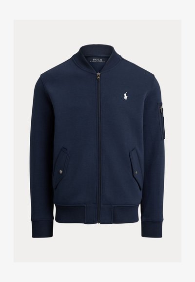 Blouson bomber bleu marine zippé avec col, poignets et bas côtelés, poches à rabat avec boutons, poche zippée sur la manche et logo Polo Ralph Lauren blanc sur la poitrine.