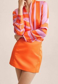 Mini-robe en satin orange vif associée à un chemisier rayé rose et orange. Le tissu a une texture lisse, avec des détails de boutons sur les manches.