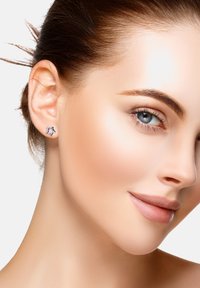 Ung kvinna med klar hy, blå ögon och ett subtilt leende, som bär stjärnformade ädelstensörhängen mot en enkel vit bakgrund.