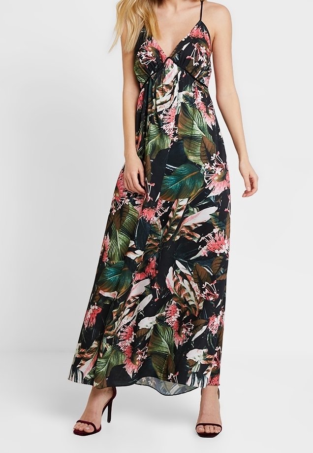 Femme portant une robe noire longue sans manches avec un imprimé floral tropical vert et rose et de fines bretelles, associée à des talons hauts noirs.