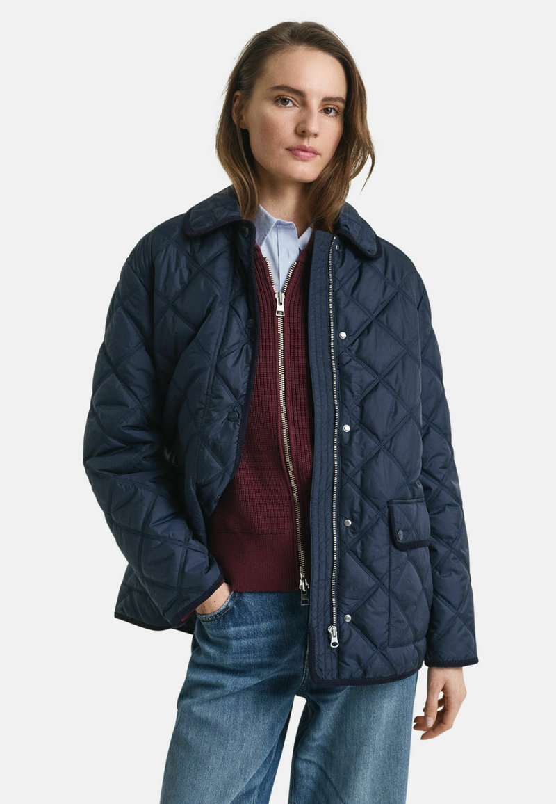 Marineblauer gesteppter Mantel mit klassischem Kragen, Frontknöpfen und einer aufgesetzten Tasche. Darunter ein bordeauxfarbener Reißverschluss-Pullover, kombiniert mit einem hellblauen Hemd und Jeans.