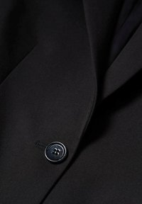 Veste en tissu noir avec une texture lisse, des revers crantés et un bouton argenté brillant avec quatre trous pour le boutonner.