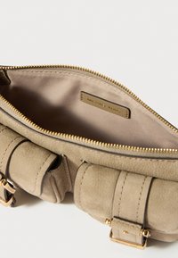 Unutrašnjost bež kožne torbe sa zatvaračem na zzip, koja sadrži oznaku brenda u zlatnoj boji i glatko platno obloge. Vidljivi akcentni hardver.