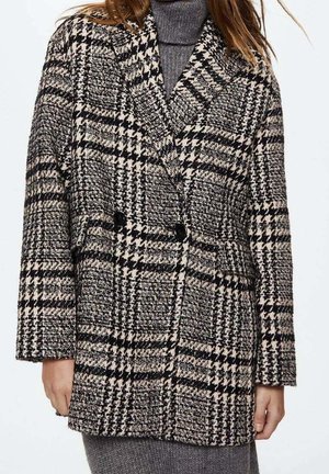 Kvinde iført en dobbeltradet frakke med sort, hvid og beige hundetandsmønster over en grå rullekrave strikket kjole.