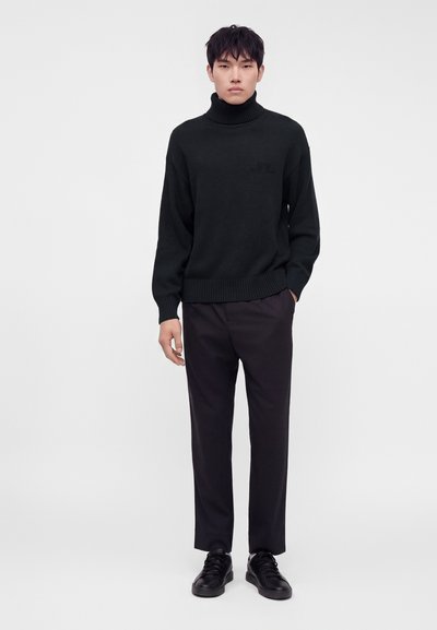 Jeune homme portant un pull noir à col roulé, un pantalon sombre et des baskets noires, debout devant un fond blanc uni.