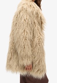 Beige shaggy fuskpälsjacka med långa, texturerade fibrer, avslappnad passform, sänka axlar och en något öppen front.
