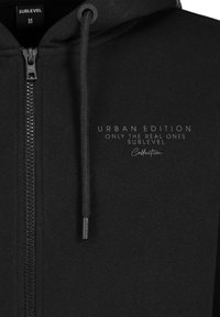 Schwarze Zip-Hoodie aus glattem Stoff mit verstellbarem Kapuzenzug, mit aufgedrucktem Logo-Text auf der Vorderseite. Standard Passform für Größe M.