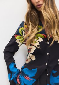 Blouse en soie noire ornée de grands motifs floraux colorés en bleu, vert et rouge, avec une patte de boutonnage sur le devant et des manches longues.