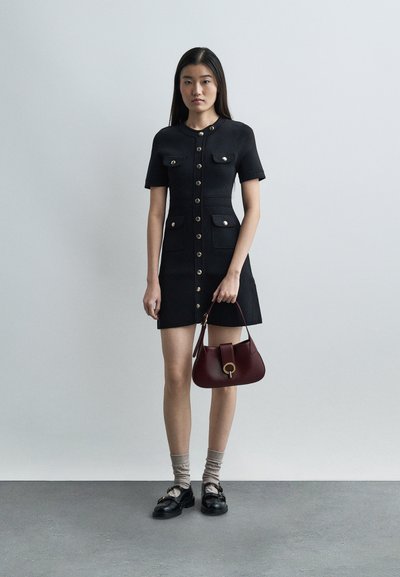 Νεαρή γυναίκα στέκεται φορώντας ένα μαύρο φόρεμα με κουμπιά μπροστά και τσέπες, μπεζ κάλτσες, μαύρα παπούτσια, κρατώντας μια μπορντό τσάντα.