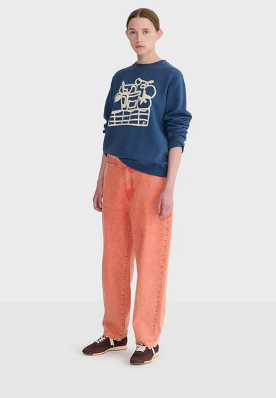 Jeune femme debout portant un sweat-shirt bleu marine avec un motif abstrait blanc, un pantalon ample orange, et des baskets foncées avec des lacets blancs.