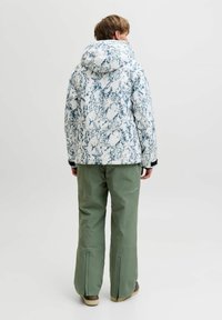 Veste blanche avec un motif marbré bleu, capuche et accents noirs ; associée à un pantalon imperméable vert et des chaussures brunes, présentée de dos.