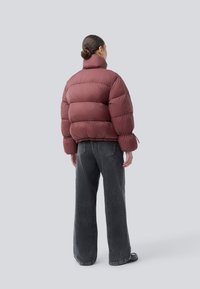 Frau mit Dutt, die eine mauvefarbene Steppjacke und weit geschnittene dunkelgraue Jeans trägt, steht mit dem Rücken zu einem schlichten grauen Hintergrund.