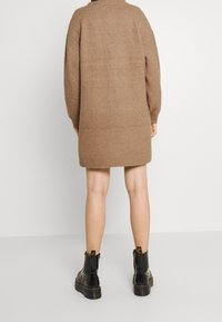 Une personne portant une robe pull camel brun longueur genoux et des bottes noires en cuir se tenant sur un sol gris clair contre un mur blanc.