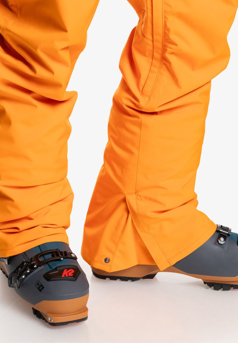 Jasnopomarańczowe spodnie narciarskie o luźnym kroju z detalami w postaci zamków błyskawicznych, w połączeniu ze szarymi i beżowymi butami narciarskimi z czarnymi klamrami i teksturowanymi podeszwami.