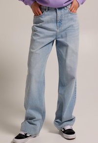 Vaqueros de mezclilla azul claro, holgados, con diseño de cinco bolsillos, costuras visibles y un desgaste sutil. Combinados con zapatillas negras.