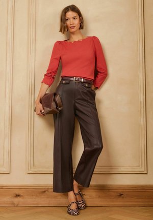 Frau trägt ein rotes Top mit geschwungenem Ausschnitt, hochgeschnittene dunkelbraune Lederhose, Leopardenmuster-Schuhe und hält eine braune Clutch.