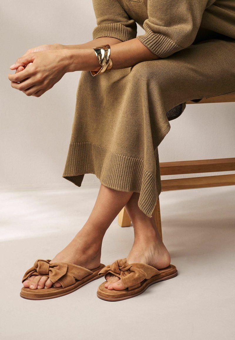 Personne assise sur un banc en bois, portant une robe en tricot marron, un bracelet manchette doré et des sandales plates en daim marron nouées en nœud sur un sol clair.