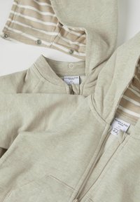 Helle beige Zip-Hoodie mit gestreiftem Innenfutter, weicher Textur, Kängurutasche und Druckknopfverschlüssen an der Kapuze.