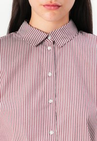 Femme portant une chemise bordeaux et blanche à rayures verticales, boutonnée avec un col pointu, visible du menton à la poitrine.