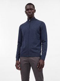 Homme portant un pull zippé bleu marine avec un col montant et un pantalon gris foncé, debout devant un fond blanc uni.