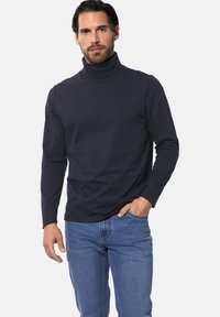 Marinefarbene Rollkragenshirt aus weichem Stoff, mit langen Ärmeln und einem dezenten Logo. Kombiniert mit blauen Jeans, die eine taillierte Silhouette zeigen.