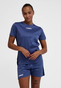 Camiseta deportiva de manga corta azul y pantalones cortos a juego con acentos laterales negros. Presenta el logo "HUMMEL" en blanco en ambas piezas. Tejido suave y transpirable.