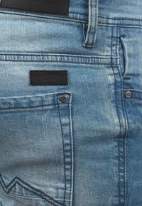 Light blue denim jeans featuring a distressed texture, a black leather patch, and double-stitched pockets with metallic rivets.