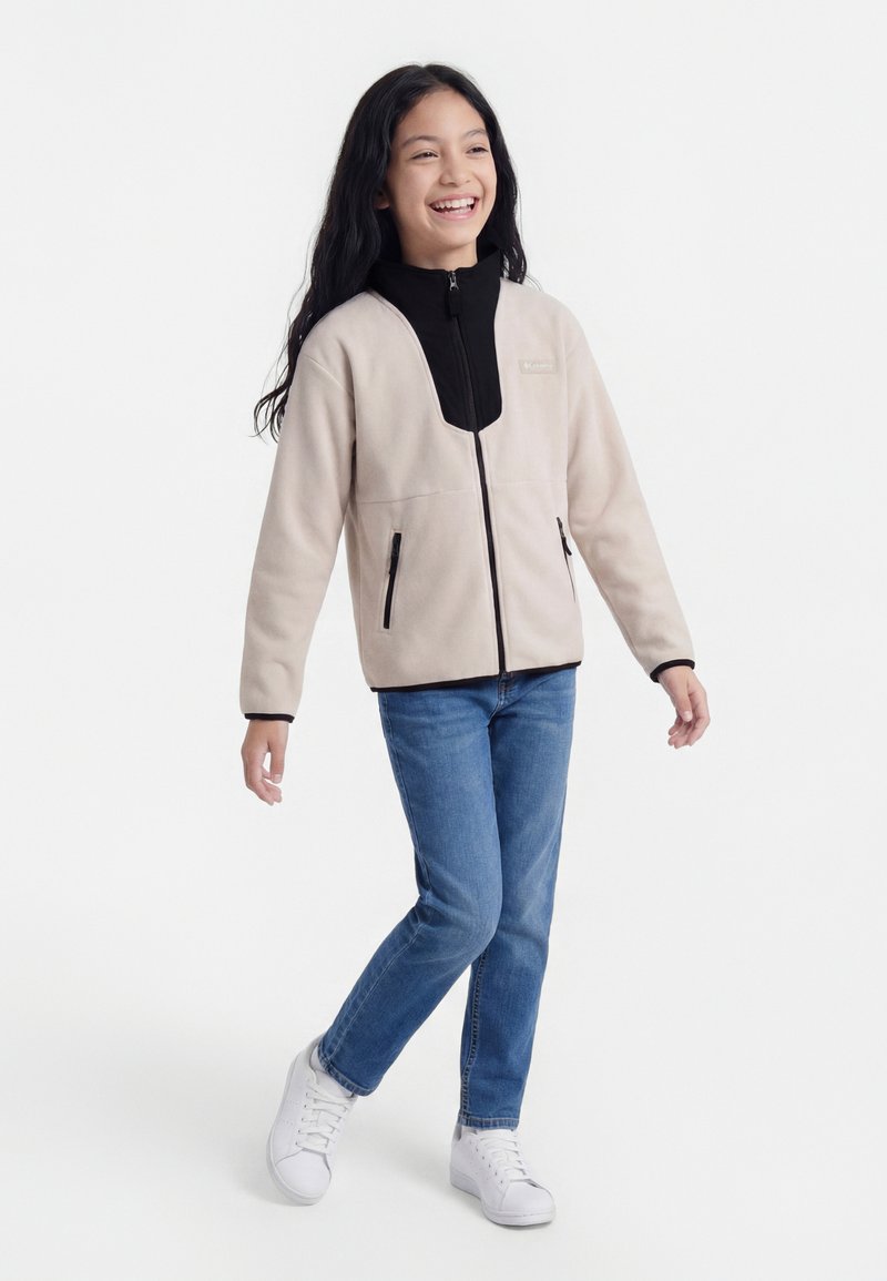 Fille souriante marchant, portant une veste beige zippée avec un col noir, un jean bleu et des baskets blanches sur fond blanc.