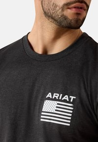 Camiseta gris oscuro con un gráfico blanco de la bandera americana y el texto "ARIAT" en letras negritas en el área izquierda del pecho.