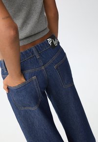 Tmavě modré džíny z denimu se rovnými nohavicemi, s oranžovým prošíváním a černou etikettou na pase. Viditelné dvě zadní kapsy.