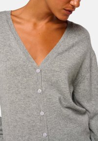 Cardigan gris avec un col en V, manches longues et six boutons lavande sur le devant. Fabriqué en tissu doux et texturé.