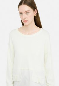 Top bianco a maniche lunghe con scollatura rotonda, caratterizzato da due tasche frontali e dettagli con cordoncino. Tessuto liscio, design a vestibilità rilassata.