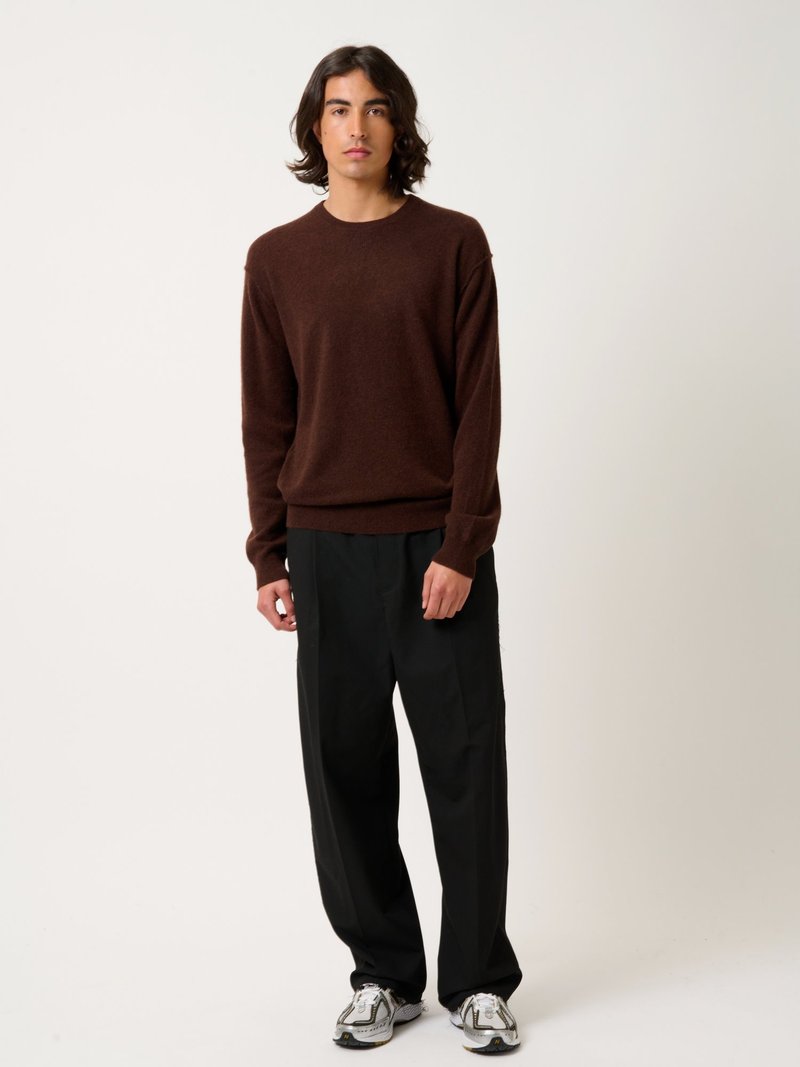 Maglione lavorato a maglia marrone, vestibilità rilassata, polsini a costine; pantaloni neri a gamba larga; sneakers bianche e grigie testurizzate. Stile minimalista, palette neutra.