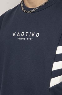Mornarska dukserica s bijelim tekstom "KAOTIKO" i natpisom "SINCE 1999" ispod. Sadrži bijele pruge na ramenima. Mekani materijal s teksturnim završetkom.