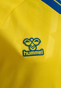 Gul sportsjakke med et struktureret mønster, en blå krave, og et blåt og gult "hummel" logo på venstre brystområde.