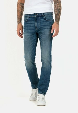 Slim-fit blue denim jeans featuring five pockets, a button and zipper closure, and subtle fading on the thighs, worn with white sneakers.