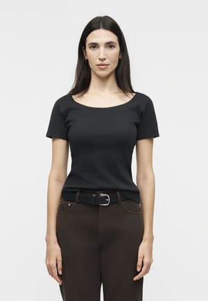 Γυναίκα με μακριά σκούρα μαλλιά, που φοράει μαύρο κοντομάνικο τοπ και σκούρο παντελόνι με μαύρη ζώνη, στέκεται μπροστά σε ένα απλό φωτεινό φόντο.