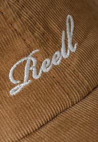 Braune Cordmütze mit weißem gesticktem Text. Der Text verläuft leicht gekrümmt und folgt der Form der Mütze. Der Stoff hat eine ausgeprägte Rippstruktur.