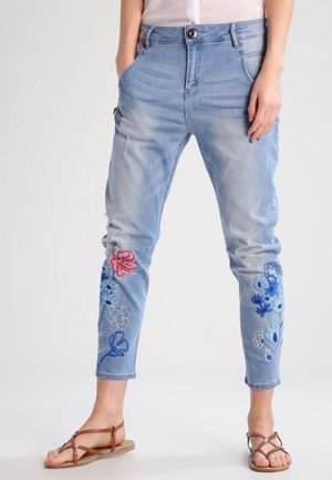 Jean court bleu clair avec broderies florales bleues et roses sur les jambes, porté avec des sandales marron à brides et une chemise blanche rentrée.