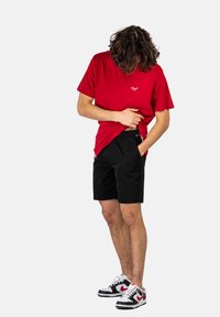 Camiseta de algodón roja con cuello redondo, pantalones cortos negros y zapatillas de deporte en blanco y negro. La camiseta presenta un pequeño logo blanco en el pecho.