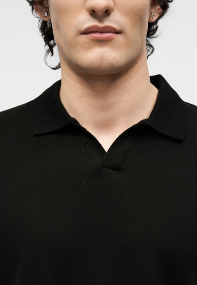 Jeune personne portant un polo noir texturé avec col, lèvres, mâchoire, menton visibles et boucles d'oreilles aux deux oreilles.