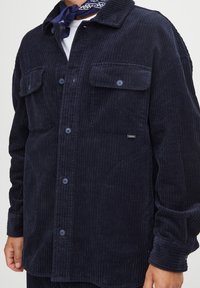 Navy corduroy shirt jacket with two front pockets, button closure, and a textured ribbed surface. Features a collared neck and buttoned cuffs.