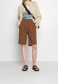 Cardigan en tricot beige, chemise à motifs floraux, shorts marron ajustés et sandales noires avec boucles dorées. Le modèle se tient contre un fond blanc.