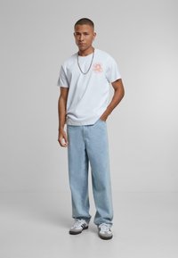 Bijela majica s kratkim rukavima i okruglim izrezom s laganim ružičastim motivom, u kombinaciji s svijetloplavim širokim trapericama i crno-bijelim tenisicama. Akcent sa srebrnim lancem.